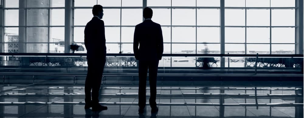 Two silhouetted figures stand facing a large window in a modern office building.