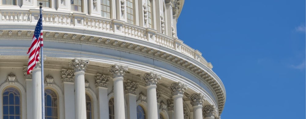 The U.S. Capitol building's dome with American flag and classical columns against a clear blue sky.