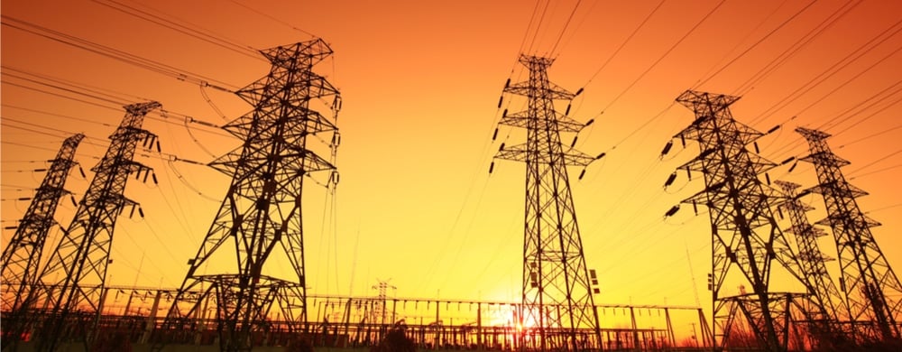Multiple high-voltage electrical transmission towers silhouetted against an orange sunset sky.