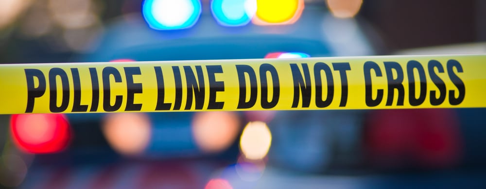 Yellow police tape with "POLICE LINE DO NOT CROSS" text in front of blurred emergency lights.